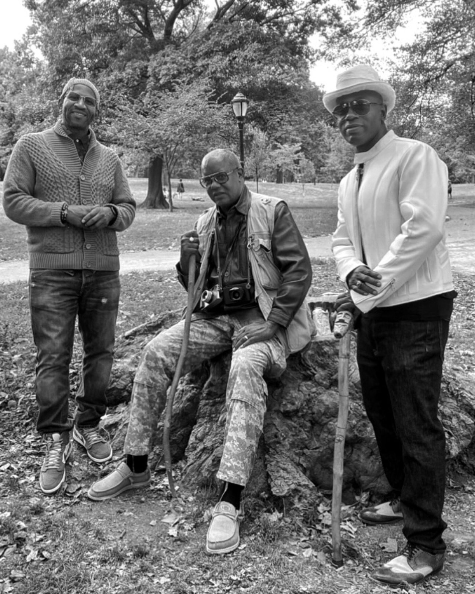 Black-and-white group photo, Prospect Park — Shabazz seated center with cameras, Murray on right, third collaborator on left